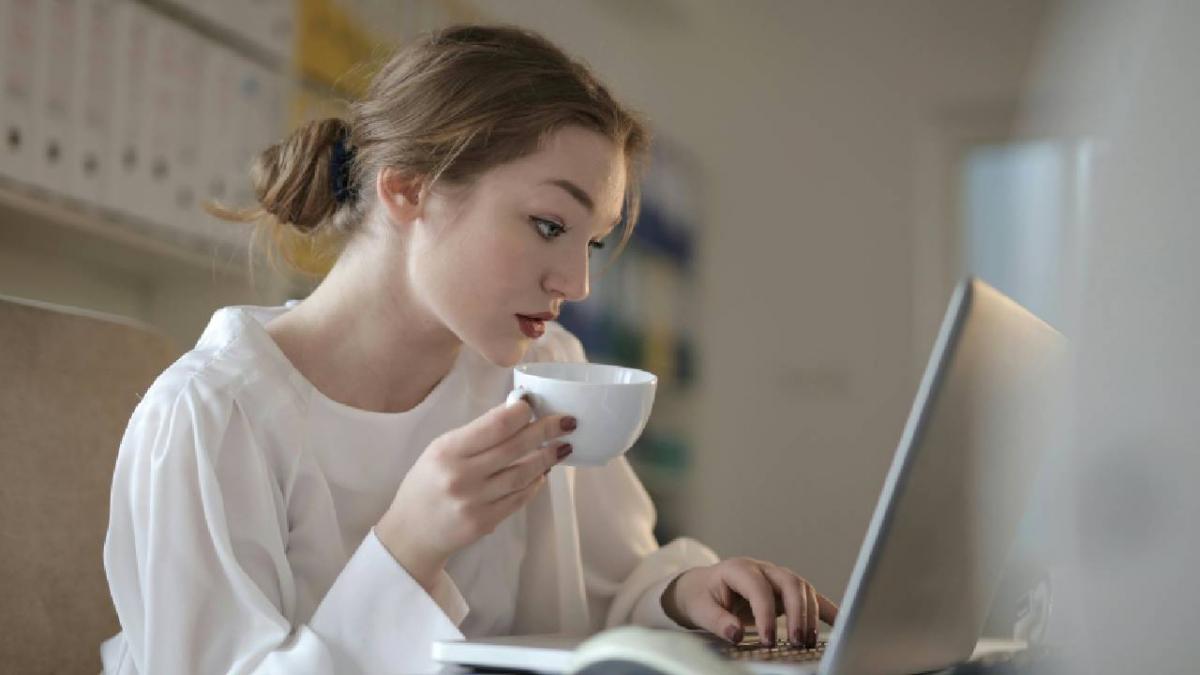 o femeie isi bea cafeaua in timp ce foloseste laptopul