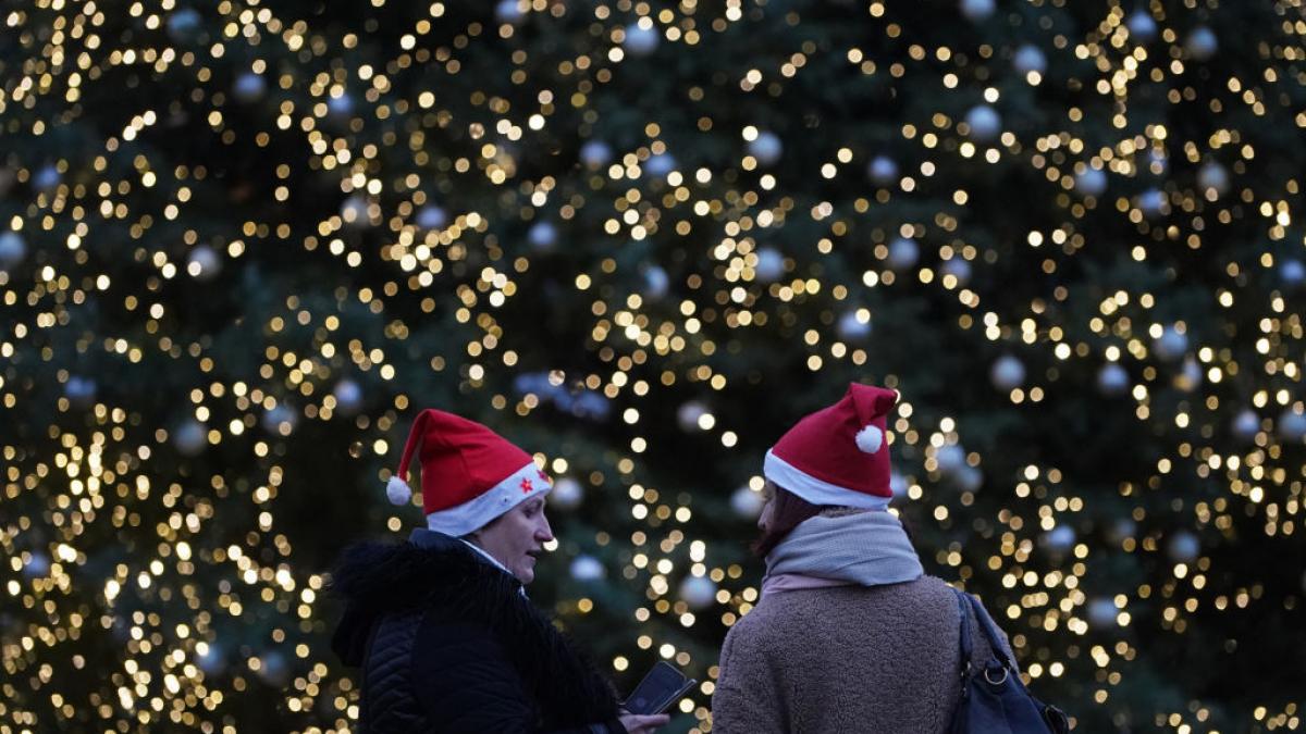 doi oameni cu căciuli de moș crăciun pe cap stau în fața unui brad