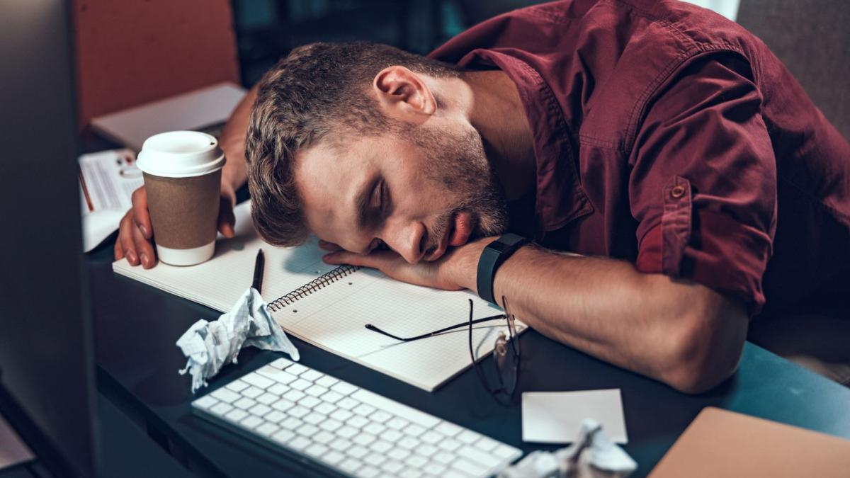 barbat care doarme cu capul pe birou și paharul de cafea in mana
