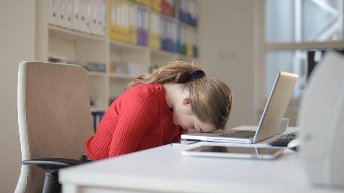 fata imbracata intr-o bluza rosie stand cu capul pe laptopul de pe birou