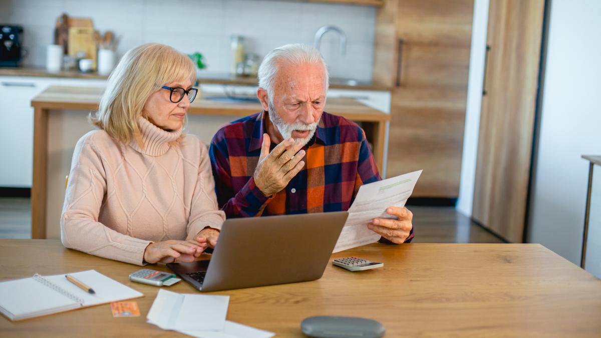 poza in care apare un cuplu de pensionari in timp ce analizeaza un document si au langa ei un laptop