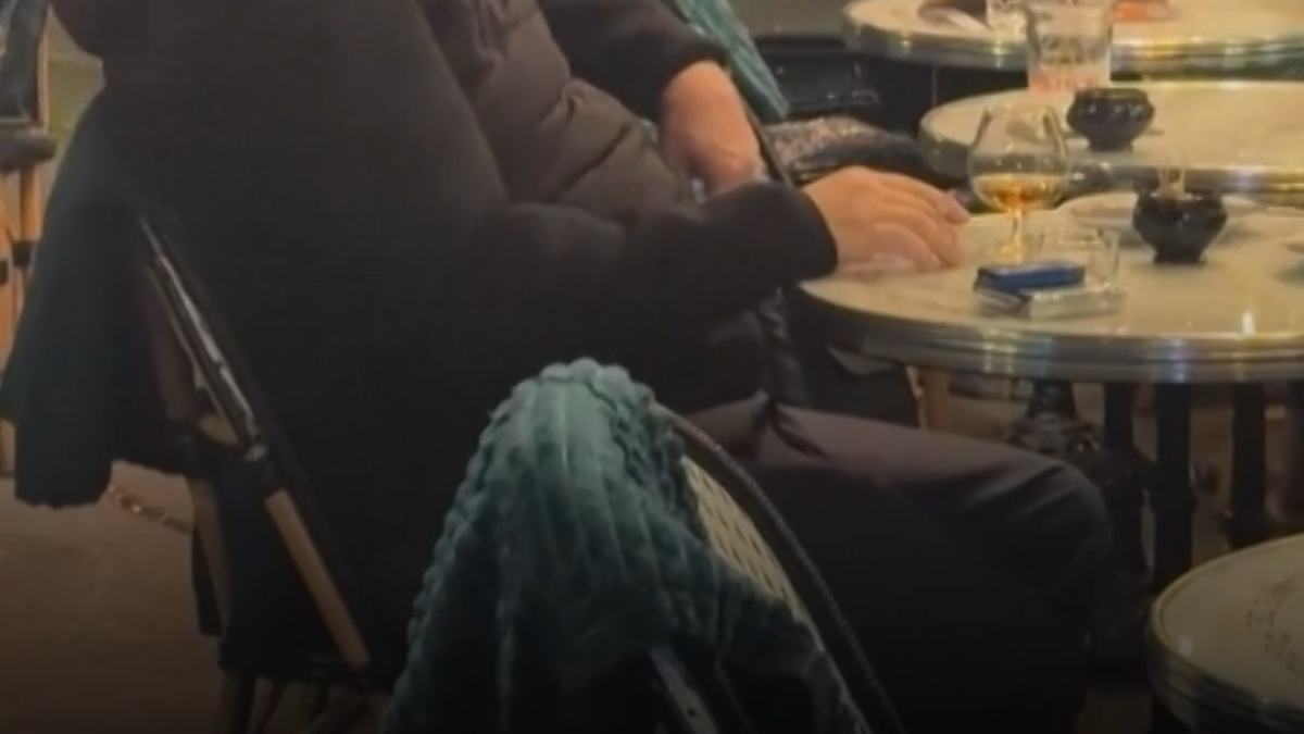 un cuplu de francezi bea vin si savureaza relaxat o tigara la o terasa in timp ce langa ei parisul arde