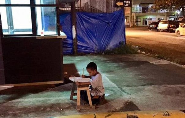 Un băieţel îşi face temele în genunchi, pe trotuar, la lumina rece a neoanelor. POVESTEA lui a devenit virală pe Facebook