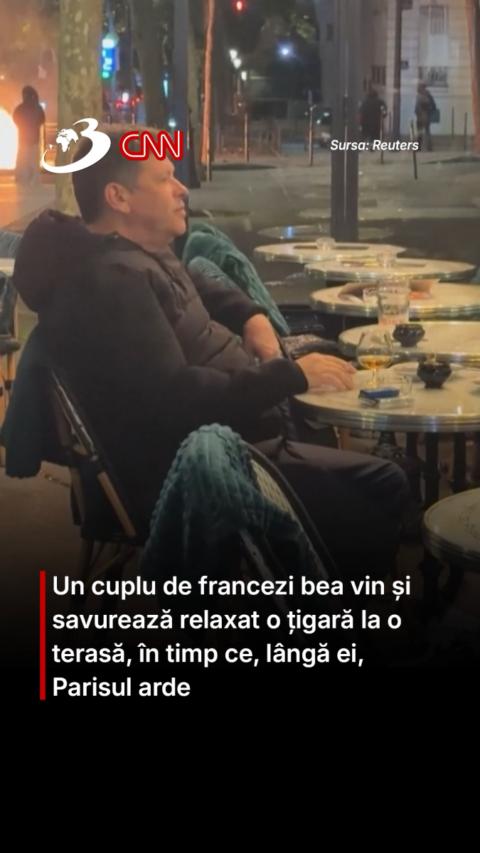 Un cuplu de francezi bea vin și savurează relaxat o țigară la o terasă, în timp ce, lângă ei, Parisul arde